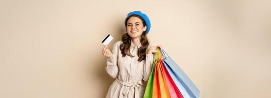 Portrat of trendy feminine girl posing with shopping bags from store and credit card, paying contactless, buying with discount on sale, beige background photo