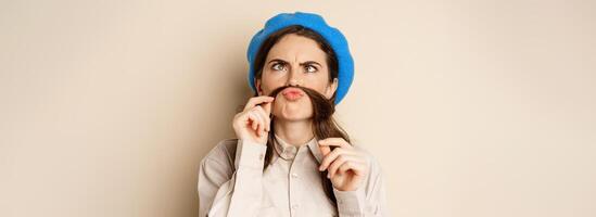 Funny beautiful girl fooling around, making moustache out of hair under lips, squinting and grimacing, standing happy against beige background photo