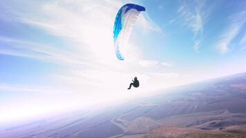 A paragliding athlete flies on a paraglider near the slope of a small mountain near a village on a sunny autumn day. Aerial view shooting with FPV drone quadcopter in acro mode video