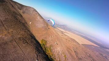 A paragliding athlete flies on a paraglider near the slope of a small mountain near a village on a sunny autumn day. Aerial view shooting with FPV drone quadcopter in acro mode video
