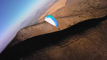 Aerial view from a sports FPV drone. A paraglider athlete flies near the slopes of a small mountain. Free paragliding flight over rural fields in autumn from FPV drone video