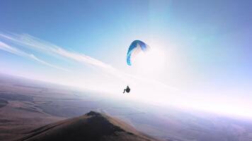 Aerial view from a sports FPV drone. A paraglider athlete flies near the slopes of a small mountain. Free paragliding flight over rural fields in autumn from FPV drone video
