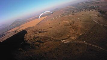 Aerial view from a sports FPV drone. A paraglider athlete flies near the slopes of a small mountain. Free paragliding flight over rural fields in autumn from FPV drone video