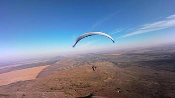 A paragliding athlete flies on a paraglider near the slope of a small mountain near a village on a sunny autumn day. Aerial view shooting with FPV drone quadcopter in acro mode video