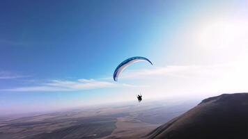 A paragliding athlete flies on a paraglider near the slope of a small mountain near a village on a sunny autumn day. Aerial view shooting with FPV drone quadcopter in acro mode video
