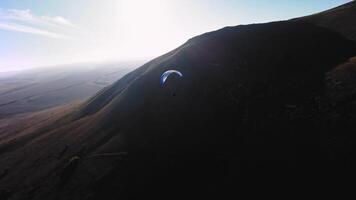 Aerial view from a sports FPV drone. A paraglider athlete flies near the slopes of a small mountain. Free paragliding flight over rural fields in autumn from FPV drone video