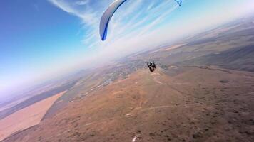 A paragliding athlete flies on a paraglider near the slope of a small mountain near a village on a sunny autumn day. Aerial view shooting with FPV drone quadcopter in acro mode video