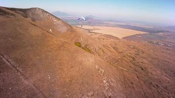 Aerial view from a sports FPV drone. A paraglider athlete flies near the slopes of a small mountain. Free paragliding flight over rural fields in autumn from FPV drone video