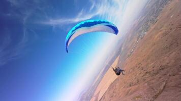 Aerial view from a sports FPV drone. A paraglider athlete flies near the slopes of a small mountain. Free paragliding flight over rural fields in autumn from FPV drone video
