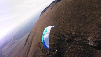 A paragliding athlete flies on a paraglider near the slope of a small mountain near a village on a sunny autumn day. Aerial view shooting with FPV drone quadcopter in acro mode video