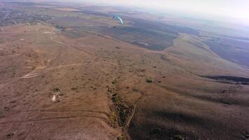 A paragliding athlete flies on a paraglider near the slope of a small mountain near a village on a sunny autumn day. Aerial view shooting with FPV drone quadcopter in acro mode video