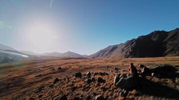 A white man wearing FPV goggles and a helmet sits in the mountains and controls an FPV acrodrone quadcopter flying around him. Dynamic footage of a drone pilot against the backdrop of epic mountains video