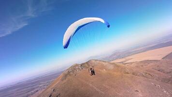 A paragliding athlete flies on a paraglider near the slope of a small mountain near a village on a sunny autumn day. Aerial view shooting with FPV drone quadcopter in acro mode video