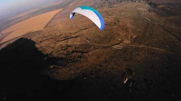 Aerial view from a sports FPV drone. A paraglider athlete flies near the slopes of a small mountain. Free paragliding flight over rural fields in autumn from FPV drone video