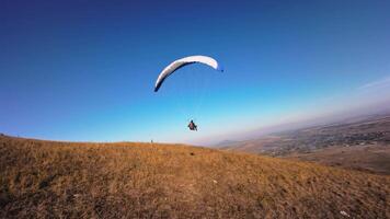 Aerial view from a sports FPV drone. A paraglider athlete flies near the slopes of a small mountain. Free paragliding flight over rural fields in autumn from FPV drone video