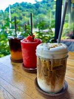 A selection of refreshing drinks including iced coffee, watermelon smoothie, and citrus iced tea on a wooden table, set against a lush tropical backdrop. Perfect for food and beverage themes. photo