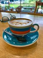 A close-up of a creamy cappuccino served in a vibrant striped cup with a matching saucer and spoon, set on a wooden table in a cozy cafe, with sunglasses and a scenic view in the background photo