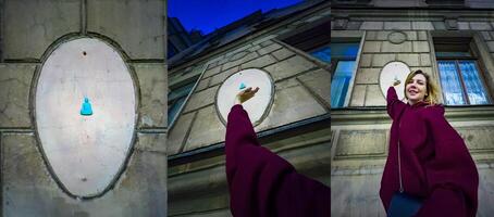 A cheerful woman in a maroon sweater reaches up towards a small blue Buddha figurine attached to the wall of a historic building. The playful interaction with the unique street art adds a touch of whimsy to the urban scene photo