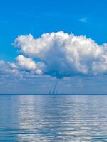 Serene view of a calm bay under a bright blue sky with large puffy clouds, featuring a distant industrial skyline on the horizon, blending the tranquility of nature with modern industry. photo