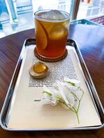Refreshing iced lemon tea served in a glass with ice cubes and lemon slices, placed on a metal tray with an open book page and white flowers, set in a cozy cafe, perfect for a relaxing afternoon. photo