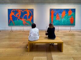 Two visitors sit on a bench in a modern art museum, closely observing the vibrant and iconic paintings by Henri Matisse, Dance and Music, creating an atmosphere of contemplation and appreciation photo