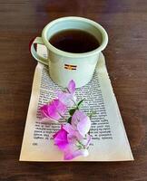 A serene setting featuring a vintage enamel cup of tea placed on an open book page, adorned with delicate pink flowers, creating a peaceful and charming ambiance on a wooden table, perfect for relaxation photo