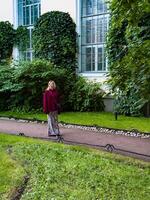 A woman in a maroon sweater and animal print skirt walks through a lush green park. The backdrop of large windows and ivy-covered walls adds to the serene and fashionable atmosphere of the scene photo