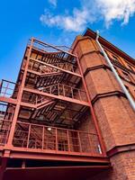 An upward view of a red metal fire escape attached to an industrial brick building. The intricate structure and contrasting colors create a visually appealing urban scene, highlighting industrial architecture photo