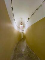 A narrow, curved hallway illuminated by vintage red chandeliers hanging from a vaulted ceiling. The hallway features beige walls and stone tiles, leading to a closed white door, creating a mysterious and historical ambiance photo
