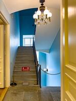 A vintage staircase illuminated by a classic chandelier in a historic building. The staircase features stone steps, ornate iron railings, and blue walls, creating an elegant and timeless interior space photo