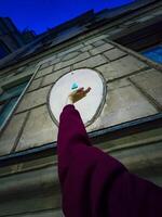 A cheerful woman in a maroon sweater reaches up towards a small blue Buddha figurine attached to the wall of a historic building. The playful interaction with the unique street art adds a touch of whimsy to the urban scene photo