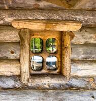 Close-up of a rustic wooden window on a log cabin, reflecting the surrounding greenery. The weathered wood and quaint window design create a charming and nostalgic scene, perfect for themes of rustic living, countryside, and vintage architecture. photo