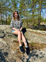 A young woman sitting on a rock in a pine forest under a clear blue sky, enjoying the serenity of nature. Perfect for themes of outdoor adventure, relaxation, and nature appreciation photo