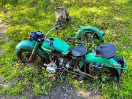An old, abandoned green motorcycle sits partially buried in overgrown grass. The weathered, rusted parts and vibrant green surroundings create a nostalgic scene, perfect for themes of decay, history, and forgotten treasures. photo