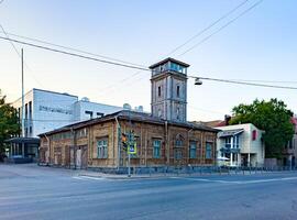 A historic wooden fire station with a distinctive tower stands at a street corner in an urban setting. The weathered structure contrasts with the modern buildings around it, highlighting the blend of old and new architecture in the city. photo