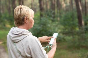 middle-aged woman in the forest takes daily steps, looks at the route map on her smartphone, turns on the pedometer, white screen mockup soft focus photo