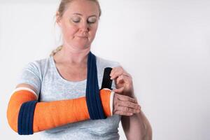 middle aged woman with broken arm in fibreglass orange cast, hangs her arm in a sling, modern treatment methods, on a neutral background, copy space photo