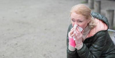 A woman with a cold blows her nose into a disposable paper handkerchief while sitting on a park bench, Her arm is broken, fixed with a modern fiberglass cast,decreased immunity due to stress photo