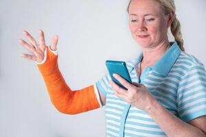 woman with broken right arm in an orange fiberglass cast doing different exercises for rehabilitation, watching a on a smartphone, modern treatment methods, on a neutral background photo