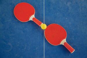 Red table tennis rackets with a yellow ball between them on a blue background, ping pong sports equipment, symmetry concept regarding the center of the table with a white line, top view, photo