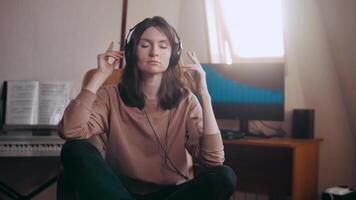 A beautiful girl with big headphones listens to music in a chair at the computer, dances, looks at the camera and smiles, the equalizer is playing on the monitor. Shakes his head to the rhythm video