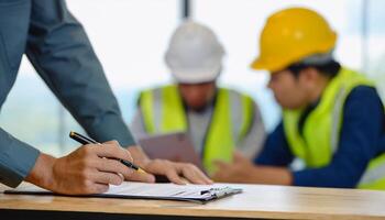 Image of Contraction site manager holding black pen and placing his hand on top of personal safety risk assessment take booklet on the table with defocused co-teamwork at the background. photo