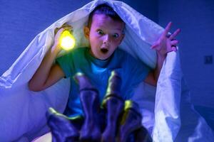 Scared boy with flashlight hides under blanket from the hand of scary monster photo