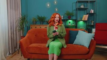 Cheerful redhead girl sitting on sofa, using smartphone share messages on social media application video