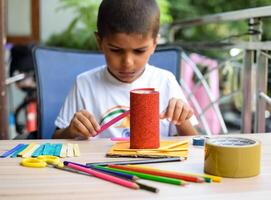 Smart Indian little boy performing best out of waste activity with different colourful objects during the summer vacations, Cute Indian Kid doing best our of waste activity at home on wooden table photo