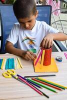 Smart Indian little boy performing best out of waste activity with different colourful objects during the summer vacations, Cute Indian Kid doing best our of waste activity at home on wooden table photo