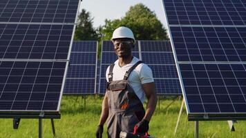 Handsome african american male mechanic in uniform standing in front of rows of solar panels holding tools and looking at camera video