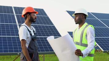 Black engineer and indian worker talking with each other while holding a blue print with a large solar panel in background video