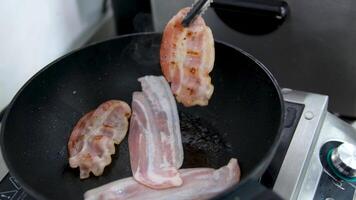 Frying pork bacon in pan side view close up slow motion. Fried slices of aromatic bacon are turned over in frying pan for better cooking. Start day with traditional breakfast. Aroma of bacon frying video