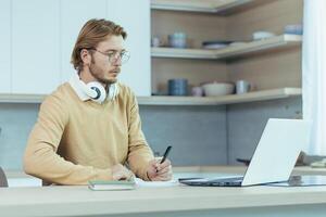 Young serious and focused man studying remotely at home, student in kitchen wearing glasses using laptop for distance learning taking notes on study material. photo